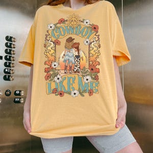 May include: A yellow short-sleeve t-shirt featuring a graphic print of two figures in cowboy hats and boots standing together. The design includes floral borders, a sun, and the text "COWBOY LIKE ME" in a light blue, stylized font.