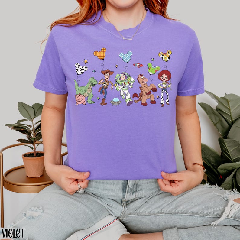 Puede incluir: Una camiseta violeta de manga corta con un estampado gr&aacute;fico de personajes de Toy Story, incluyendo a Woody, Buzz Lightyear, Jessie, Rex, Bullseye y Hamm. Los personajes sostienen globos coloridos con forma de orejas de rat&oacute;n y estrellas.