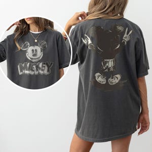 May include: A charcoal gray short-sleeve t-shirt featuring a distressed graphic of Mickey Mouse on the front and back. The front displays a sketch-style Mickey face with the text Mickey below it, while the back shows a full-body rear view of Mickey.