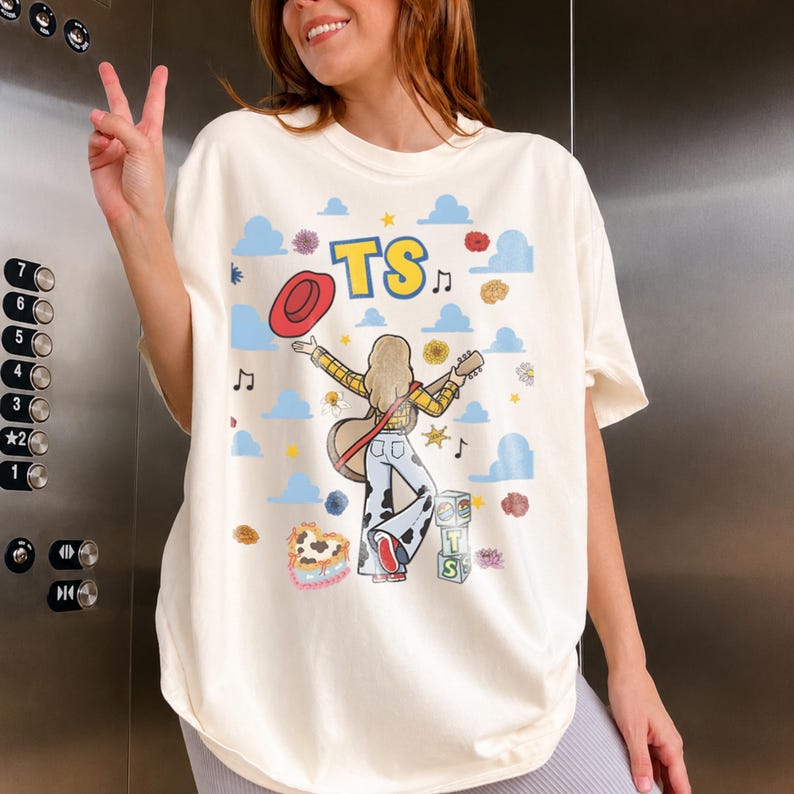 May include: An off-white t-shirt featuring a graphic print of a person with blonde hair wearing a yellow plaid shirt and cow-print pants, holding a guitar and a red hat. The design includes blue clouds, musical notes, flowers, and blocks with the text TS.