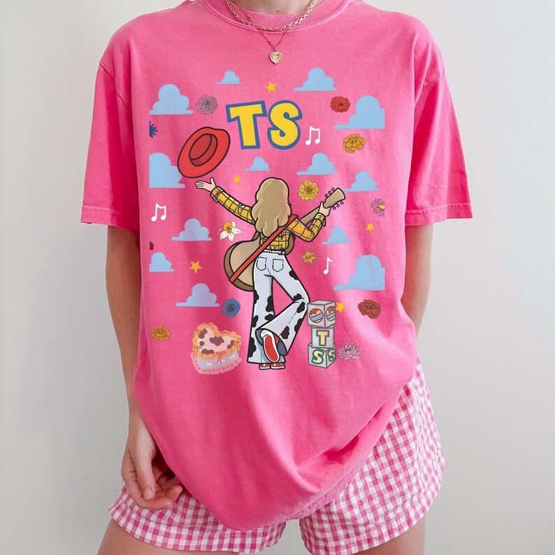 May include: A bright pink t-shirt featuring a graphic of a person with blonde hair playing an acoustic guitar, wearing a yellow plaid shirt and cow-print pants. The design includes the text TS, a red hat, blue clouds, flowers, and blocks.