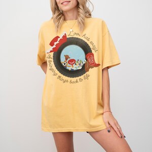 May include: A yellow short-sleeve t-shirt featuring a graphic of a black tire framing a scene with cartoon characters, a red cowboy hat, and a red cowboy boot. The text reads, Love has ways of bringing things back to life, in brown script.