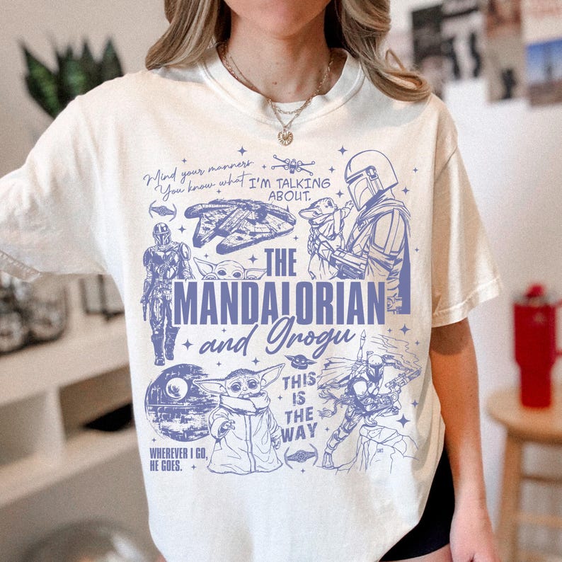 May include: A cream-colored short-sleeve t-shirt featuring a blue graphic print of The Mandalorian and Grogu. The design includes various Star Wars characters, spaceships, and the text "The Mandalorian and Grogu," "This is the way," and "Mind your manners."