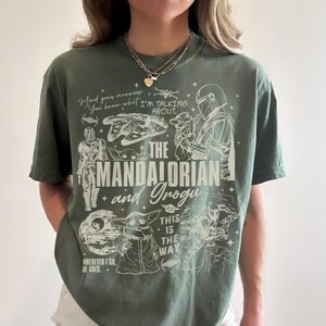 May include: An olive green short-sleeve t-shirt featuring a white graphic print of The Mandalorian and Grogu. The design includes the text "The Mandalorian and Grogu," "Mind your manners," "I'm talking about," and "This is the way," along with illustrations of the Millennium Falcon, the Death Star, and various characters.