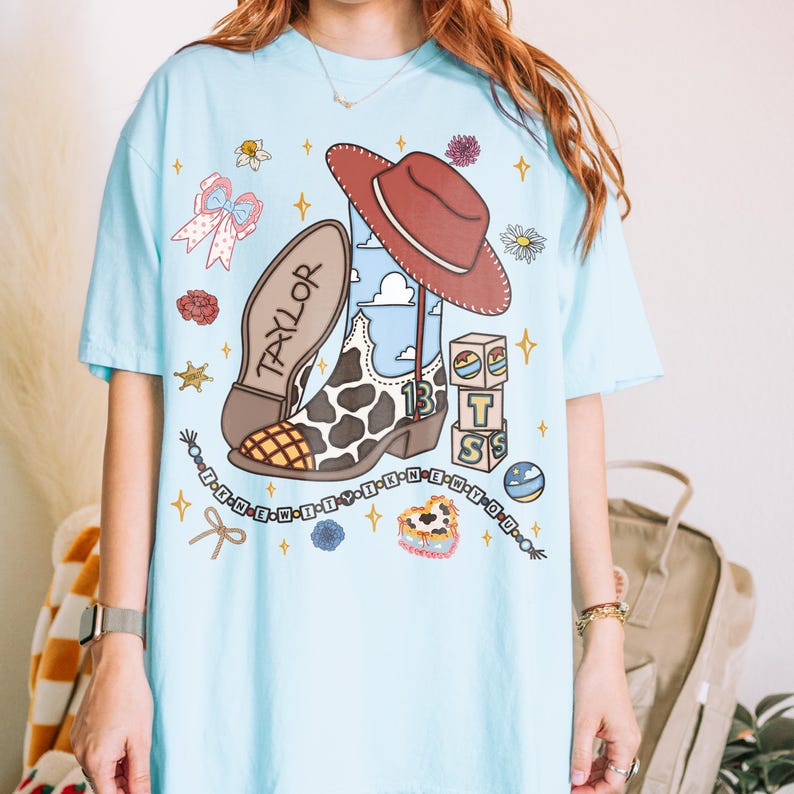 Puede incluir: Una camiseta azul claro con un estampado gr&aacute;fico de una bota vaquera con patr&oacute;n de manchas de vaca, un sombrero marr&oacute;n, bloques de letras que forman BTS, un collar de cuentas con el texto I KNEW IT Y I KNEW YOU y varios iconos de flores y estrellas.