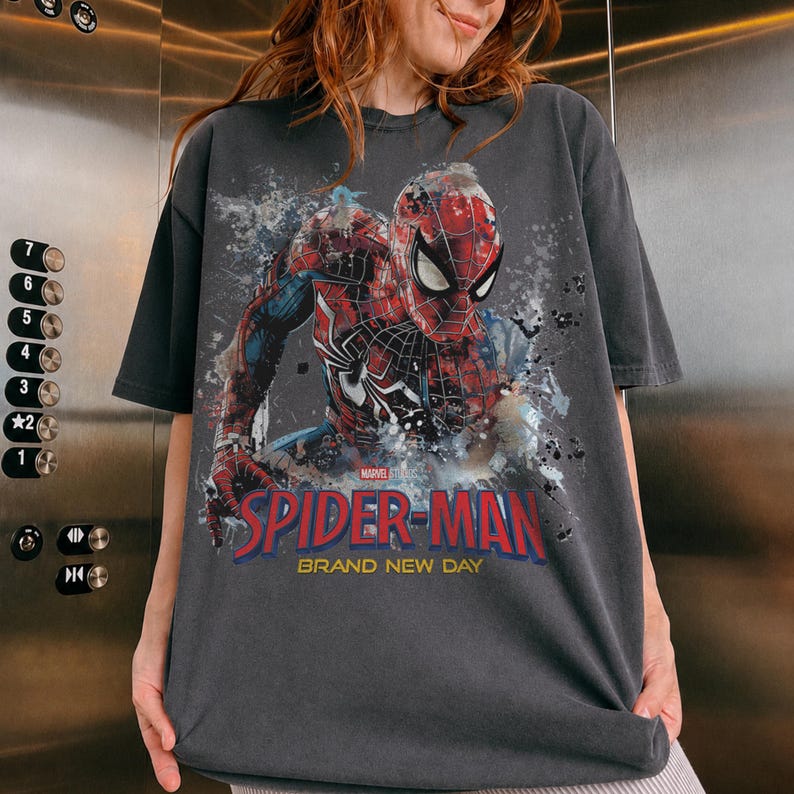 May include: A charcoal gray short-sleeve t-shirt featuring a graphic of Spider-Man in a red and blue suit. The design includes paint splatter effects, the Marvel logo, and the text "SPIDER-MAN BRAND NEW DAY" printed in bold red and yellow letters.