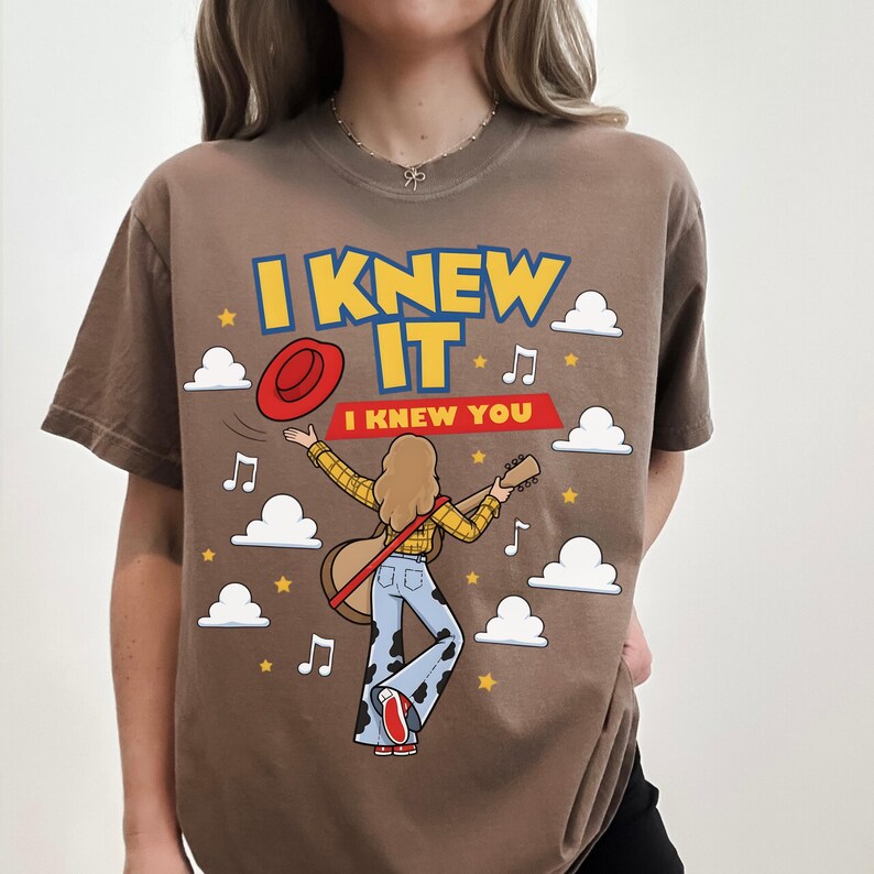 May include: A brown t-shirt featuring a graphic of a person with long hair playing a guitar, wearing a yellow plaid shirt and blue cow-print bell-bottom pants. The design includes a red hat, white clouds, yellow stars, musical notes, and the text I KNEW IT I KNEW YOU.