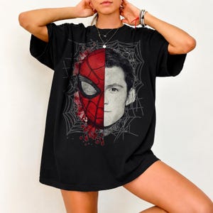 May include: A black short-sleeve t-shirt featuring a split-face graphic design. One half displays a red and black Spider-Man mask, while the other half shows a realistic portrait of a young man, all set against a grey spiderweb background.