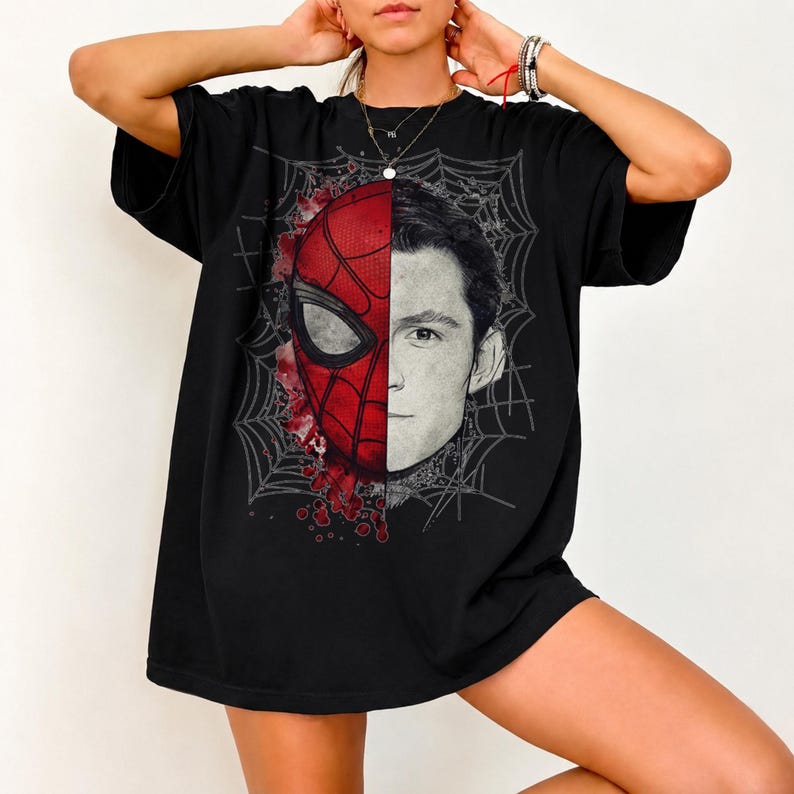 May include: A black short-sleeve t-shirt featuring a split-face graphic design. One half displays a red and black Spider-Man mask, while the other half shows a realistic portrait of a young man, all set against a grey spiderweb background.