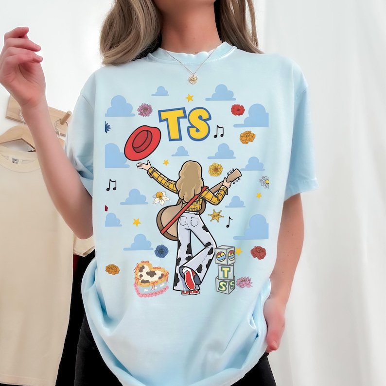 May include: A light blue t-shirt featuring a graphic of a person with blonde hair wearing a yellow plaid shirt and cow-print pants, holding a guitar and a red hat. The design includes the text TS, blue clouds, musical notes, and flowers.