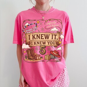 May include: A bright pink short-sleeve t-shirt featuring a graphic print with a brown scroll banner that reads I KNEW IT, I KNEW YOU. The design includes a cowboy hat, boots, alphabet blocks, a lasso, stars, and the text infinity and beyond.