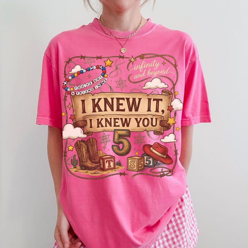 May include: A bright pink short-sleeve t-shirt featuring a graphic print with a brown scroll banner that reads I KNEW IT, I KNEW YOU. The design includes a cowboy hat, boots, alphabet blocks, a lasso, stars, and the text infinity and beyond.