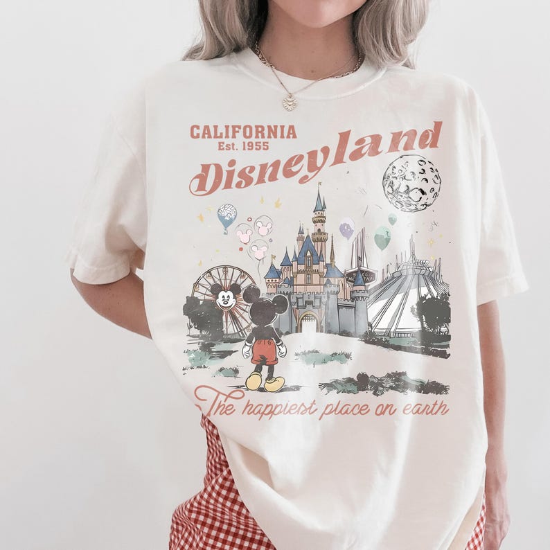 May include: Cream-colored t-shirt featuring a Disneyland graphic. The design includes the text "CALIFORNIA Disneyland Est. 1955" with an illustration of Mickey Mouse, a castle, and other park elements. The phrase "The happiest place on earth" is also included.