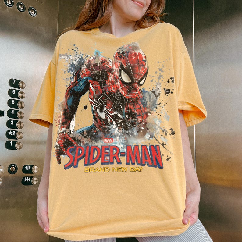 May include: A mustard yellow short-sleeve t-shirt featuring a graphic of Spider-Man in a red and blue suit. The design includes paint splatter effects, the text "MARVEL STUDIOS", "SPIDER-MAN" in large red letters, and "BRAND NEW DAY" in yellow.