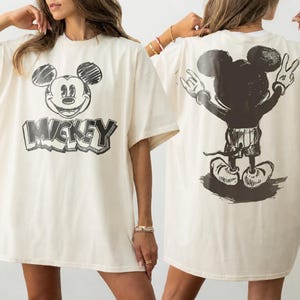May include: An off-white short-sleeve t-shirt featuring a black sketch-style graphic of Mickey Mouse's face and the text MICKEY on the front, and a black sketch-style graphic of Mickey Mouse from behind with hands raised on the back of the shirt.