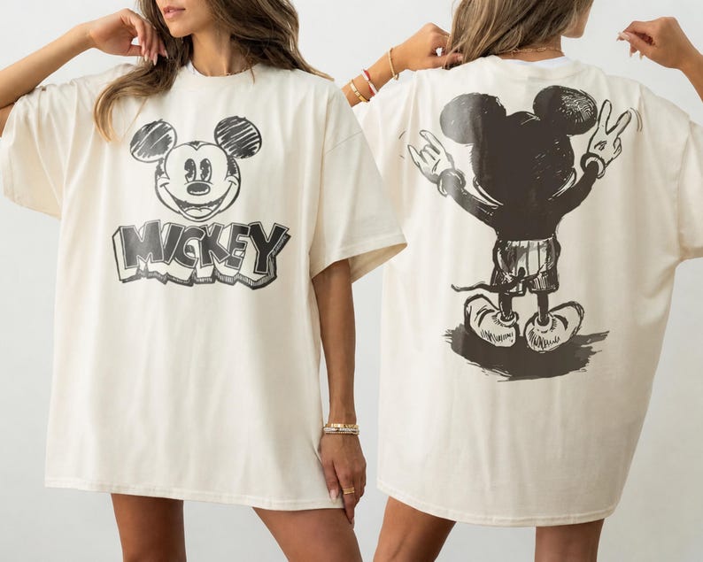 May include: An off-white short-sleeve t-shirt featuring a black sketch-style graphic of Mickey Mouse's face and the text MICKEY on the front, and a black sketch-style graphic of Mickey Mouse from behind with hands raised on the back of the shirt.