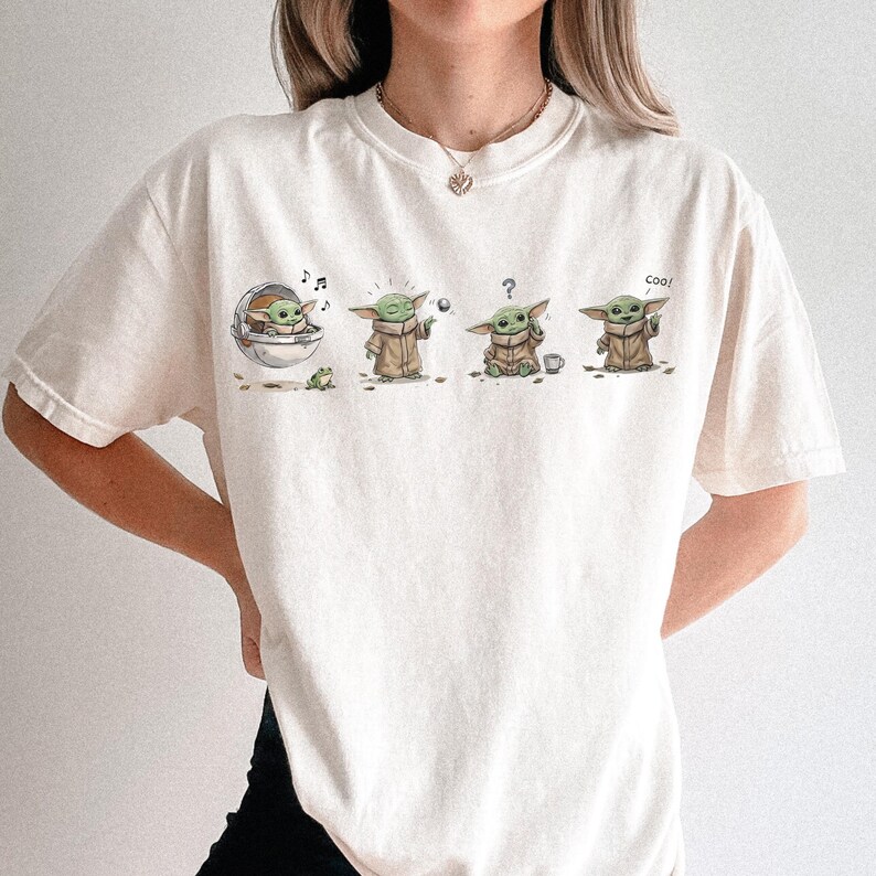 May include: An off-white short-sleeve t-shirt featuring a graphic print of a small green creature in four different poses. The illustrations show the character in a floating pod, holding a ball, looking confused with a question mark, and waving.