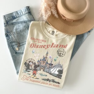 May include: Cream-colored Disneyland t-shirt with a graphic of Mickey Mouse, a castle, and the text "California Disneyland Est. 1955". The shirt is paired with light blue jeans, a gold necklace, and a tan hat.