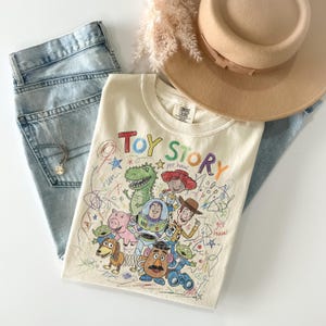 Peut inclure: Un t-shirt cr&egrave;me avec un graphisme color&eacute; "Toy Story" avec divers personnages et le texte "Toy Story". Le t-shirt est associ&eacute; &agrave; un jean bleu clair, un chapeau beige et un collier dor&eacute;.