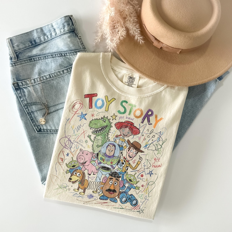 Peut inclure: Un t-shirt cr&egrave;me avec un graphisme color&eacute; "Toy Story" avec divers personnages et le texte "Toy Story". Le t-shirt est associ&eacute; &agrave; un jean bleu clair, un chapeau beige et un collier dor&eacute;.