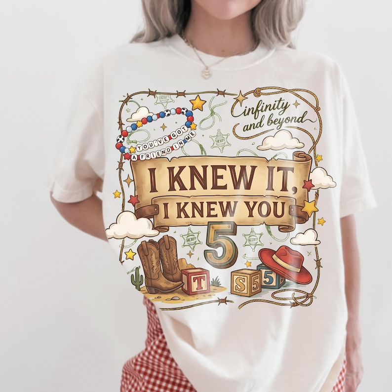 May include: A cream-colored t-shirt featuring a graphic print with a scroll banner that reads "I KNEW IT, I KNEW YOU" and "5" in the center. The design includes cowboy boots, a red hat, alphabet blocks, a beaded bracelet, and the text "Cinfinity and beyond".