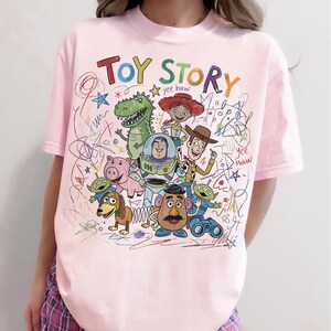 Peut inclure: Un t-shirt rose clair avec un graphisme color&eacute; des personnages du film d'animation "Toy Story". Le design comprend les mots "Toy Story" dans une police ludique, ainsi que des images de Woody, Buzz l'&Eacute;clair et d'autres personnages.