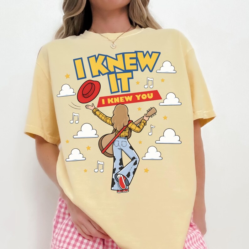 May include: A pale yellow t-shirt featuring a graphic of a person with long hair playing an acoustic guitar, wearing a yellow plaid shirt and blue cow-print bell-bottom pants. The text reads I KNEW IT and I KNEW YOU, surrounded by clouds, stars, and musical notes.