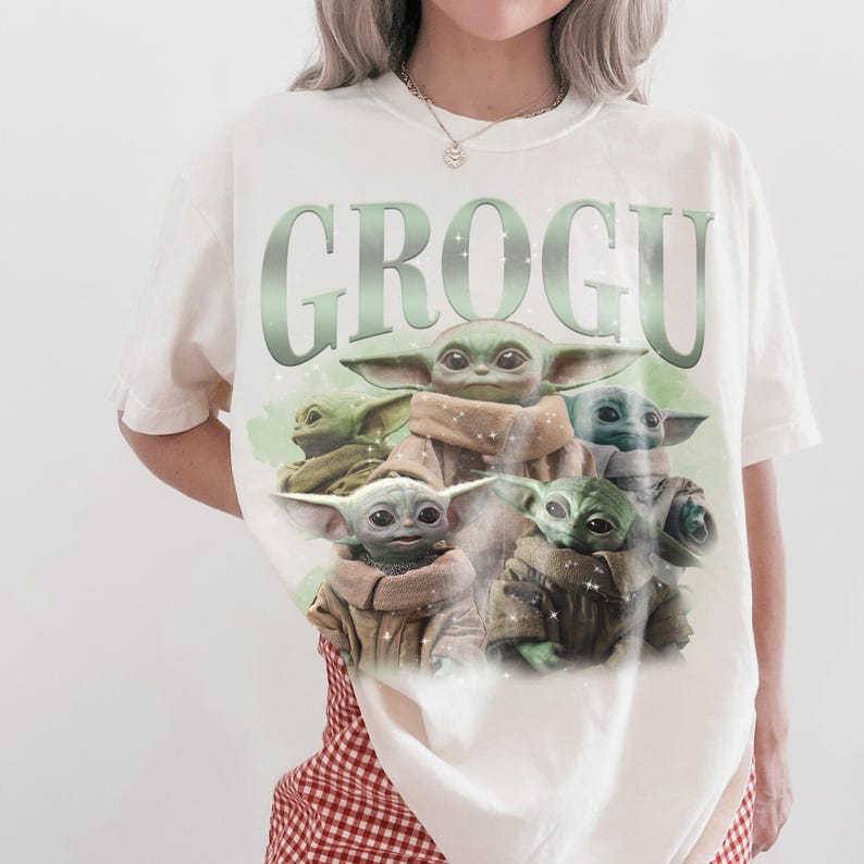May include: A cream-colored short-sleeve t-shirt featuring a large graphic print of multiple green alien creatures with large ears and eyes. The text GROGU is printed in large, light green letters above the characters, which are wearing tan robes.