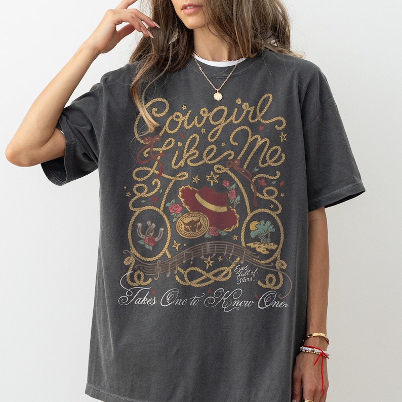 Pu&ograve; includere: Una t-shirt a maniche corte grigio antracite con una stampa grafica che riporta il testo Cowgirl Like Me e Takes One to Know One. Il design include un cappello rosso, un ferro di cavallo, un bordo a corda, stelle e piccoli dettagli floreali.