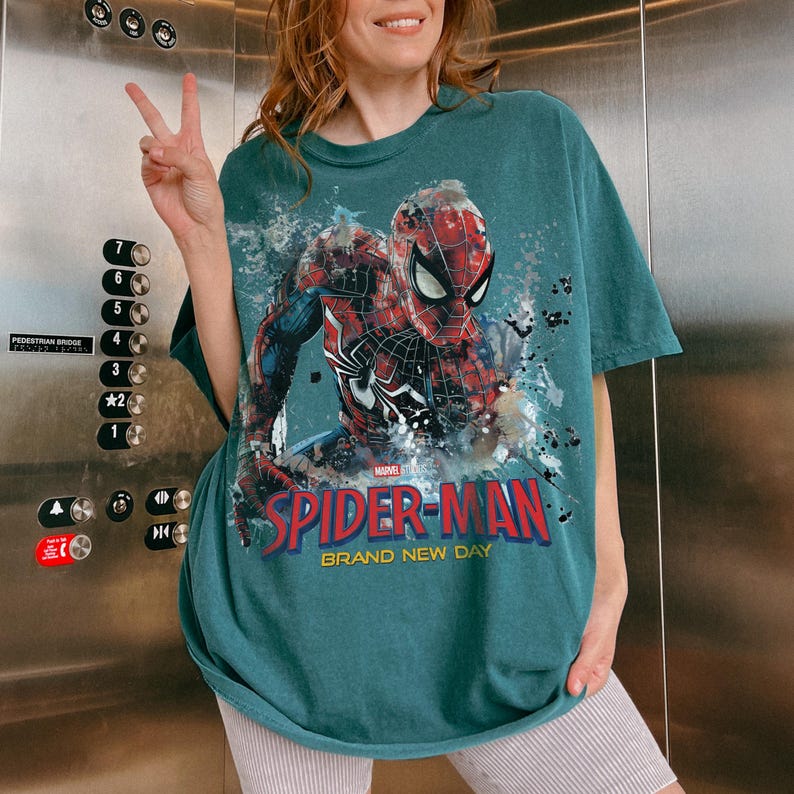 May include: A teal short-sleeve t-shirt featuring a large graphic of the Marvel character Spider-Man in a dynamic pose. The design includes paint splatter effects, the red and blue Spider-Man logo, and the text Spider-Man Brand New Day.