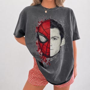 May include: A charcoal gray short-sleeve t-shirt featuring a split-face graphic print. The left side displays a red and black Spider-Man mask, while the right side shows a black and white portrait of a man, all set against a spiderweb background.