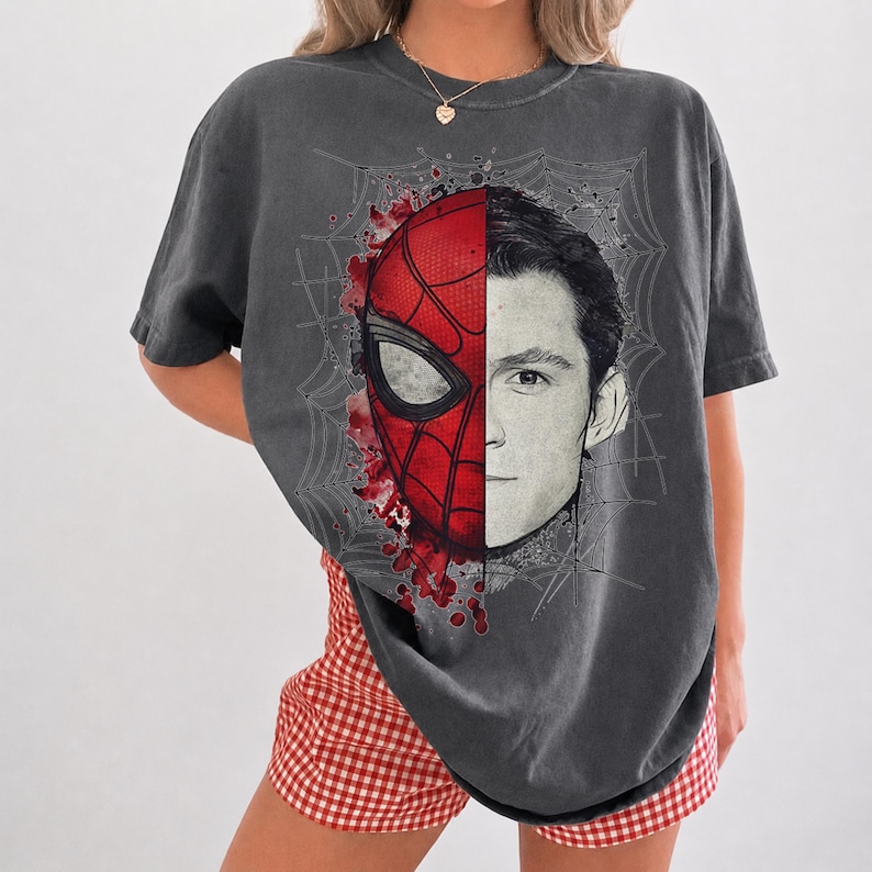 May include: A charcoal gray short-sleeve t-shirt featuring a split-face graphic print. The left side displays a red and black Spider-Man mask, while the right side shows a black and white portrait of a man, all set against a spiderweb background.