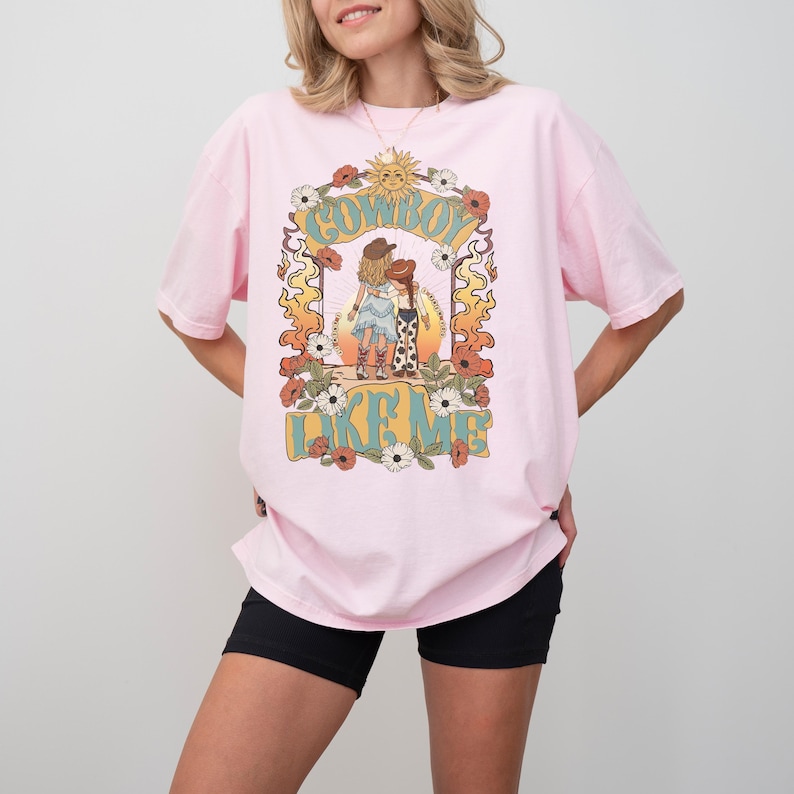 May include: A light pink t-shirt featuring a graphic print of two figures in cowboy hats standing together. The design includes floral elements, a sun, and the text "COWBOY LIKE ME" in a stylized font. The shirt has short sleeves and a crew neckline.