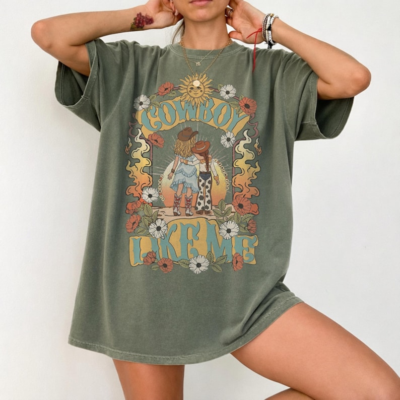 May include: An olive green short-sleeve t-shirt featuring a graphic print of two figures in cowboy hats standing before a sun. The text "COWBOY LIKE ME" is printed in blue letters, surrounded by a decorative border of orange flames and flowers.