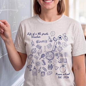 Puede incluir: Una camiseta de manga corta en color gris jaspeado claro con un estampado gráfico en azul oscuro de varios iconos escolares, incluyendo un globo terráqueo, libros y una manzana. El texto dice "Life of a 7th grade Teacher" y "Name school Est. 2026".