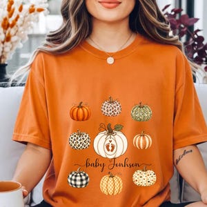 May include: An orange short-sleeve t-shirt featuring a graphic print of various pumpkins in different patterns and colours, including leopard print, plaid, and stripes. The central pumpkin displays a pair of baby footprints and the text Baby Jonhson.