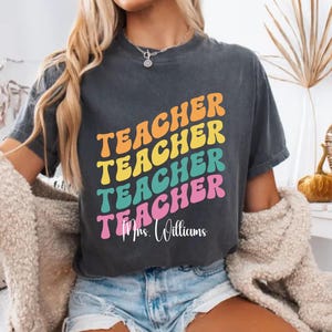 Puede incluir: Una camiseta gris carbón de manga corta con la palabra TEACHER repetida cuatro veces en una fuente retro ondulada y apilada en naranja, amarillo, verde azulado y rosa. Debajo del texto, el nombre Mrs. Williams está impreso en cursiva blanca.