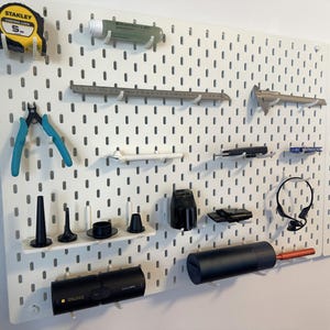 May include: A white pegboard displaying various tools and accessories. Items include a yellow tape measure, blue pliers, a ruler, a caliper, a flashlight, and other small black and white objects. The pegboard is mounted on a white wall.
