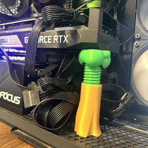 May include: A 3D-printed green and gold computer graphics card support bracket. The bracket is designed to provide support for a graphics card inside a computer case. The bracket has a screw-like design and is made of plastic.
