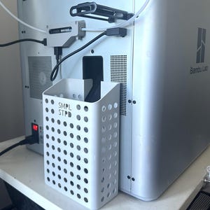 Könnte beinhalten: Ein weißer, perforierter rechteckiger Halter mit dem Text "SMPL STPD" ist an einem silbernen 3D-Drucker befestigt. Der Drucker hat das Logo "Bambu Lab" auf der Seite. Kabel und andere Komponenten sind sichtbar.