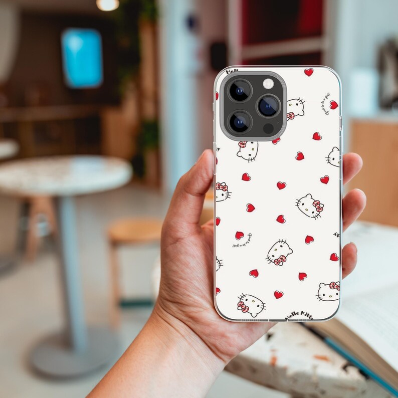 Puede incluir: Una funda de tel&eacute;fono blanca con un patr&oacute;n de caras de Hello Kitty y corazones rojos. La funda tiene un borde transparente y est&aacute; sujeta en la mano de una persona. Las caras de Hello Kitty tienen lazos rosas.