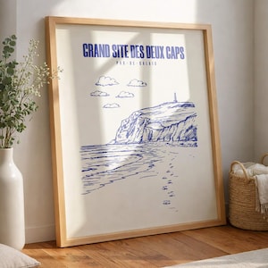 Op de afbeelding: Ingelijste kunstprint met een blauw-witte illustratie van een kustlandschap. De print bevat de tekst "GRAND SITE DES DEUX CAPS" en "PAS-DE-CALAIS". De afbeelding toont een strand, kliffen, wolken en een vuurtoren.