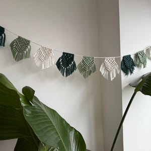 Könnte beinhalten: Eine Makramee-Girlande mit blattförmigen Designs in verschiedenen Grün- und Off-White-Tönen hängt vor einer weißen Wand. Die Girlande ist an einer dünnen weißen Schnur befestigt und bildet einen dekorativen Akzent. Eine große grüne Pflanze befindet sich im Vordergrund.