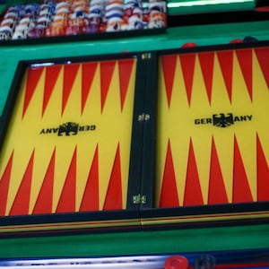 Könnte beinhalten: Ein Backgammon-Spiel mit einem gelben und roten Dreiecksmuster auf der Spielfläche. Das Wort "GERMANY" ist auf dem Brett aufgedruckt. Schwarze und rote Spielsteine ​​sind um das Brett herum verstreut, das sich auf einer grünen Oberfläche befindet.