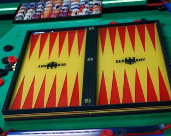 May include: A backgammon set with a yellow and red triangular pattern on the playing surface. The word "GERMANY" is printed on the board. Black and red game pieces are scattered around the board, which is set on a green surface.