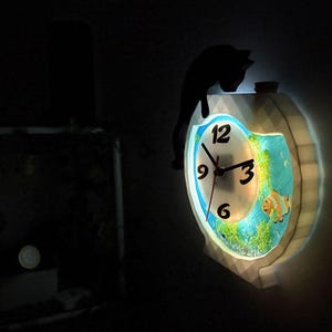 May include: A decorative wall clock with a fish tank design. The clock face has black numbers and hands. A black cat silhouette sits atop the clock. The clock is illuminated with a soft glow, showcasing the fish and aquatic plants.