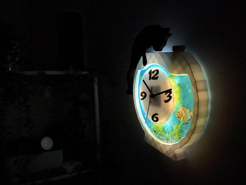 May include: A decorative wall clock with a fish tank design. The clock face has black numbers and hands. A black cat silhouette sits atop the clock. The clock is illuminated with a soft glow, showcasing the fish and aquatic plants.