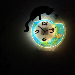 May include: A decorative wall clock with a unique design. The clock face features a fish tank scene with a clownfish and aquatic plants. A black cat silhouette sits above the clock. The clock has a glowing effect.