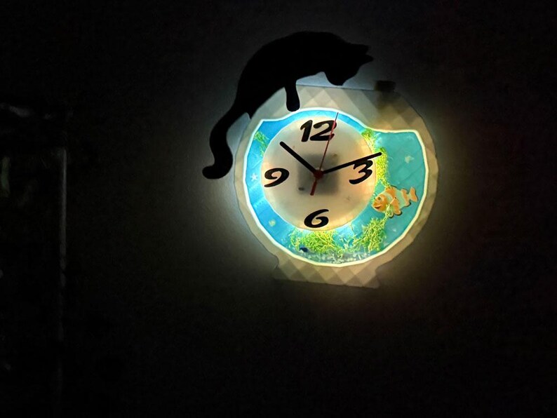 May include: A decorative wall clock with a unique design. The clock face features a fish tank scene with a clownfish and aquatic plants. A black cat silhouette sits above the clock. The clock has a glowing effect.