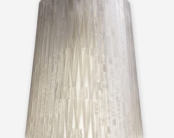 May include: A white, textured lampshade with a tapered shape. The shade has a geometric pattern of vertical and angled lines, creating a layered effect. The lampshade is suspended from a white cord.